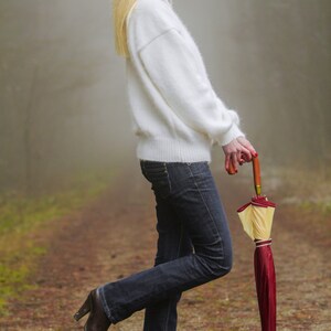 May include: A woman in a white fuzzy sweater and blue jeans is holding a red and yellow umbrella. She is standing on a path in a foggy forest.