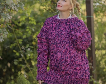 May include: A chunky knit sweater in a vibrant pink and navy blue speckled pattern. The oversized sweater has long sleeves and a crew neck. The model is wearing a white collared shirt underneath. The background is a lush green garden.