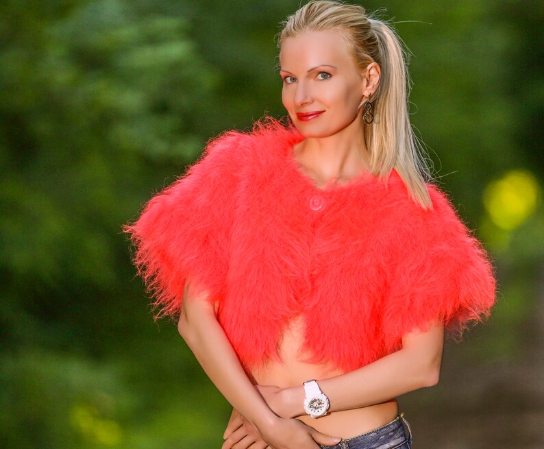 May include: A vibrant coral bolero jacket with a fluffy texture and a single button. The cropped jacket is made of a soft material. The background is a blurred green, suggesting an outdoor setting.