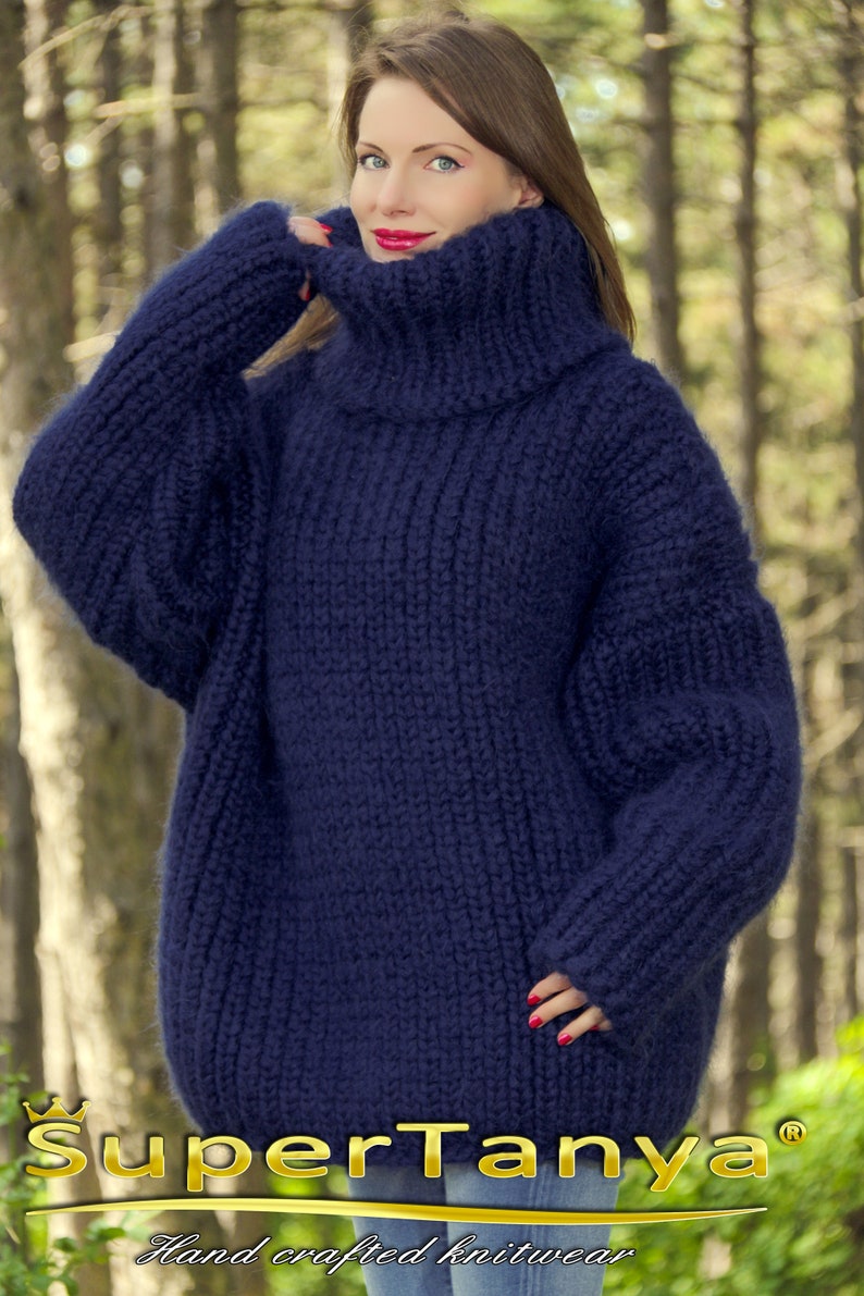 May include: A navy blue oversized turtleneck sweater with a loose fit and long sleeves. The sweater is made of a soft, fuzzy yarn and has a relaxed, comfortable look. The sweater is hand-knitted and features a ribbed texture. The text "SuperTanya&reg; Hand crafted knitwear" is visible at the bottom of the image.