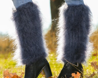 May include: A pair of dark gray leg warmers made of fluffy material, worn over blue jeans and black ankle boots with block heels. The boots are on a grassy surface with fallen leaves, suggesting an autumn setting.