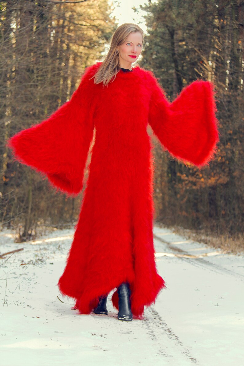 May include: A full-length, vibrant red, fuzzy coat with wide bell sleeves. The coat is worn over black boots in a snowy, wooded environment. The coat's texture appears soft and plush.