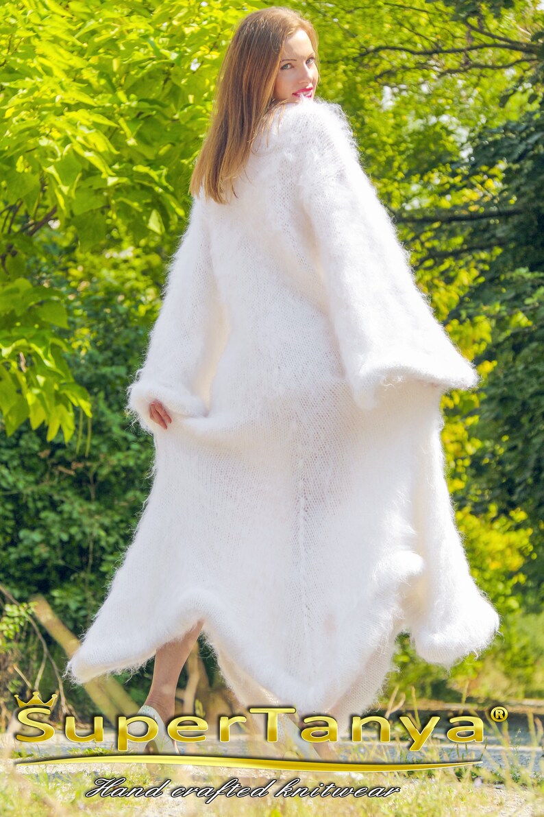 May include: A long, white, fluffy knit cardigan with wide sleeves. The cardigan is floor-length and appears soft and warm. The image includes the text "SuperTanya" and "Hand crafted knitwear". The background is a lush green outdoor setting.