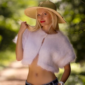 May include: A woman wearing a white, fluffy, short-sleeved cardigan and a wide-brimmed straw hat with a red accent. The cardigan is buttoned at the top. The background is a blurred green, suggesting an outdoor setting.