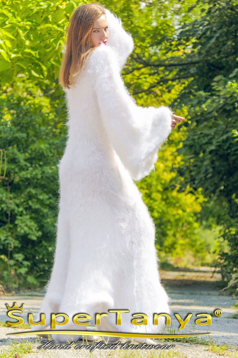 May include: A long, white, fluffy knit dress with wide sleeves. The dress is floor-length and appears soft and warm. The brand name "SuperTanya" is visible in gold lettering at the bottom of the image, along with the text "Hand crafted knitwear".