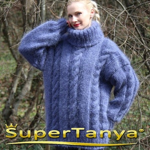 Könnte beinhalten: Ein blauer, übergroßer Zopfstrickpullover mit hohem Rollkragen. Der Pullover hat lange Ärmel und ein strukturiertes, flauschiges Aussehen. Der Text "SuperTanya" wird in goldfarbenen Buchstaben angezeigt.