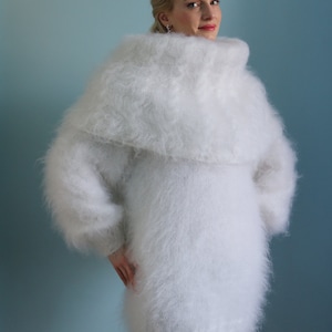 May include: A fluffy, white, oversized sweater with a wide, draped collar. The sweater has long sleeves and a soft, textured appearance. The model is wearing the sweater against a light blue background.