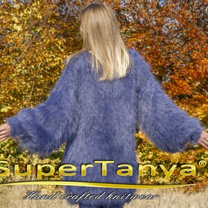 May include: A long-sleeved, fluffy, periwinkle blue knit sweater. The sweater has a loose fit and a soft, textured appearance. The brand name "SuperTanya" and the text "Hand crafted knitwear" are visible at the bottom of the image.