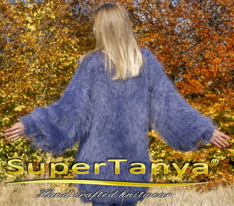 May include: A long-sleeved, fluffy, periwinkle blue knit sweater. The sweater has a loose fit and a soft, textured appearance. The brand name "SuperTanya" and the text "Hand crafted knitwear" are visible at the bottom of the image.