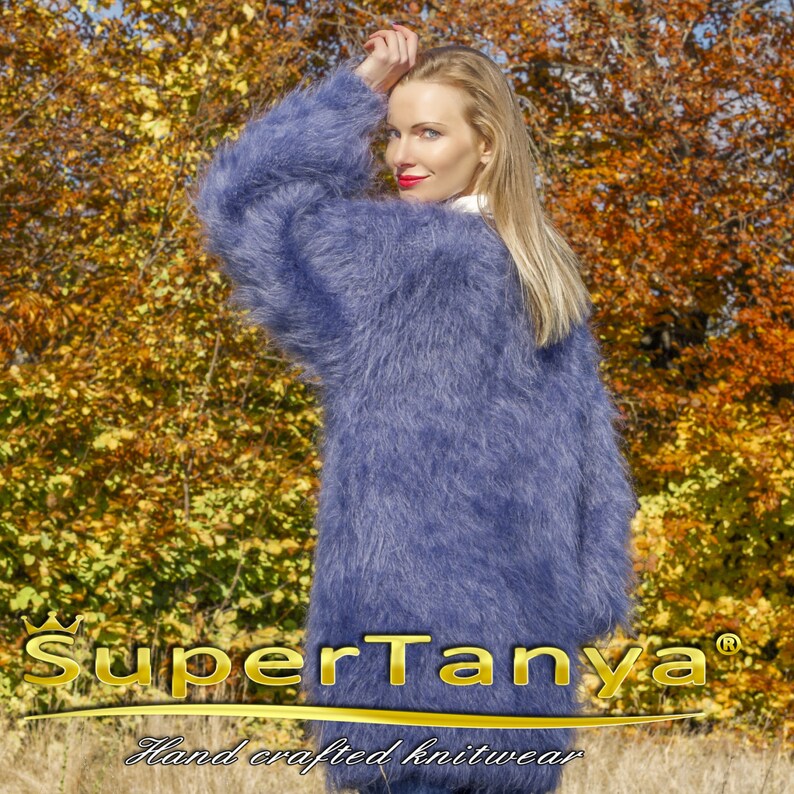 May include: A long, fluffy, periwinkle blue knit cardigan. The cardigan is worn by a person with long blonde hair and red lipstick. The background features fall foliage. The text "SuperTanya" and "Hand crafted knitwear" are visible.