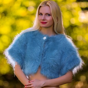 May include: A light blue, fluffy, knitted shrug with a single white button fastening. The short-sleeved bolero-style sweater is made of a soft, fuzzy material. The background is a blurred view of green and yellow foliage.