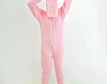 May include: A light pink knitted onesie with a matching hood that covers the head and face, featuring eye and mouth cutouts. The onesie has long sleeves, a full-length zipper, and attached feet. The outfit is made of a soft, textured material.