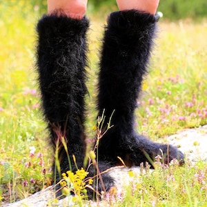 Könnte beinhalten: Ein Paar schwarze, flauschige Kniestrümpfe mit weicher, flauschiger Textur. Die Strümpfe werden von einer Person getragen, die in einem Feld mit grünem Gras und Wildblumen steht.
