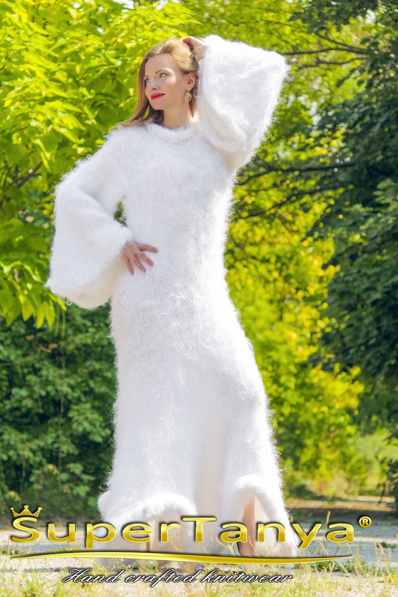 May include: A long, white, fuzzy knit dress with bell sleeves. The dress is floor-length and has a soft, textured appearance. The image includes the text "SuperTanya" and "Hand crafted knitwear".