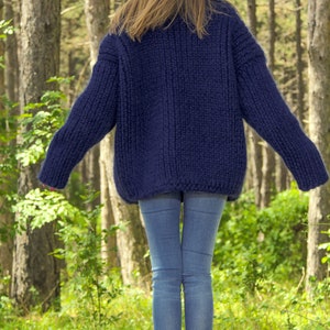 May include: A woman wearing a dark blue, chunky knit sweater and blue jeans. The sweater is hand-knitted and has a relaxed fit. The woman is standing in a forest setting. The text "SuperTanya&reg; Hand crafted knitwear" is visible at the bottom of the image.