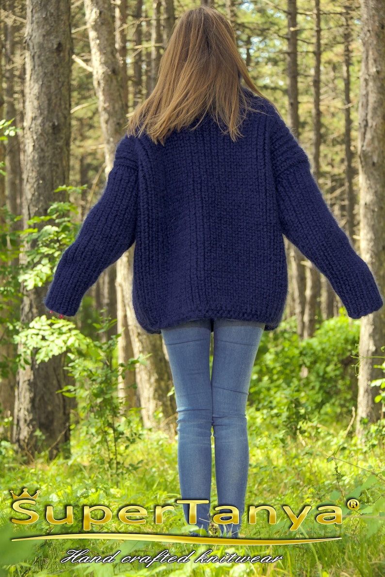 May include: A woman wearing a dark blue, chunky knit sweater and blue jeans. The sweater is hand-knitted and has a relaxed fit. The woman is standing in a forest setting. The text "SuperTanya&reg; Hand crafted knitwear" is visible at the bottom of the image.