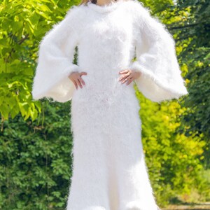 May include: A long, white, fluffy knit dress with long, flared sleeves. The dress has a high neckline and a flowing skirt. The image includes the text "SuperTanya" and "Hand crafted knitwear" in gold lettering.