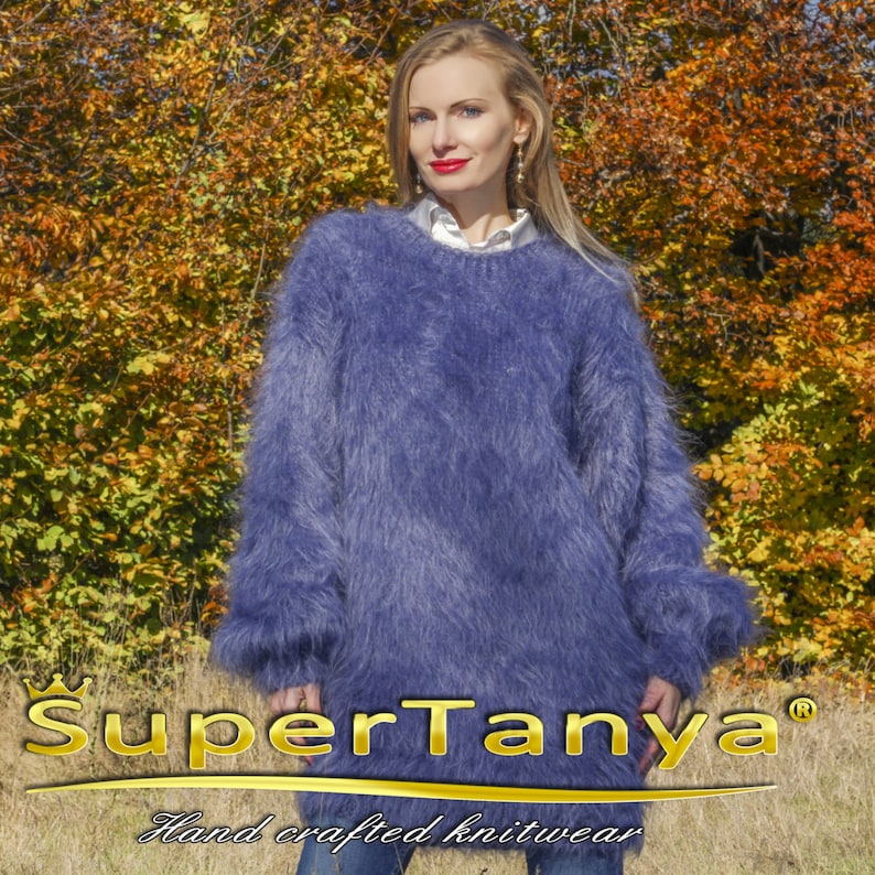 May include: A long-sleeved, oversized, fuzzy blue sweater. The sweater has a crew neck and long sleeves with a fluffy texture. The brand name "SuperTanya" and the text "Hand crafted knitwear" are displayed at the bottom of the image.