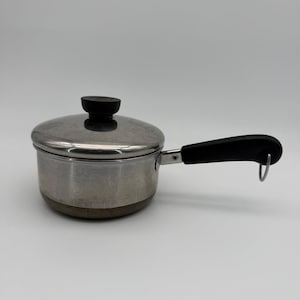 May include: A stainless steel saucepan with a matching lid, featuring a black plastic handle and a black knob on the lid. The handle includes a small metal loop at the end for hanging. The base of the pan has a copper-colored finish.