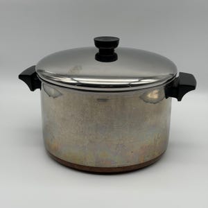 Pode incluir: Uma panela de aço inoxidável com acabamento polido e base na cor cobre. A panela possui duas alças laterais pretas e uma tampa correspondente com um botão preto no topo. A superfície externa apresenta sinais de uso e descoloração.