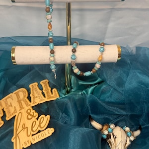 May include: Two beaded bracelets featuring turquoise, brown, and glittery silver beads, displayed on a gold-toned T-bar stand. A small animal skull adorned with matching beads sits nearby, alongside a wooden sign reading FERAL & free BY NICOLE.