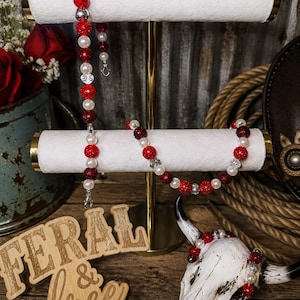 May include: A beaded accessory set featuring red glittery spheres, white pearls, and silver-toned spacers. The set includes a long strand, a shorter bracelet, and a smaller piece wrapped around a white skull. A wooden sign reads FERAL & free BY NICOLE.