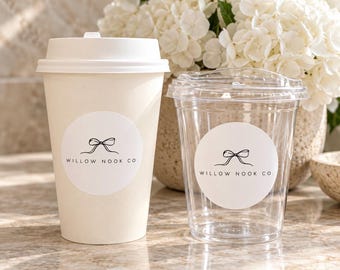 May include: A white paper cup with a lid and a clear plastic cup with a lid, both featuring a circular white sticker with a black line drawing of a bow and the text WILLOW NOOK CO. printed in black on the front of each cup.