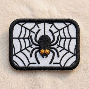 May include: A rectangular embroidered patch featuring a black spider centred on a white spiderweb background. The spider has eight legs and two prominent golden-yellow eyes. The entire patch is framed by a thick, black stitched border.