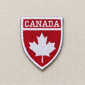 Op de afbeelding: Een schildvormige geborduurde patch met een rode achtergrond en een wit esdoornblad in het midden. Het woord CANADA is in witte hoofdletters geborduurd in een horizontale witte balk aan de bovenkant van de patch, die een witte rand heeft.