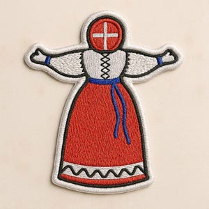 May include: An embroidered patch shaped like a doll with a red circular head featuring a white cross. The figure wears a white blouse with black lace-up detailing, a long red skirt with a white zigzag hem, and a blue waist tie with dangling ends.