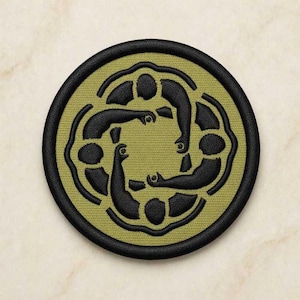 Puede incluir: Un parche bordado circular con fondo verde oliva y borde negro. El diseño muestra cuatro figuras negras estilizadas dispuestas en formación circular, con los brazos extendidos hacia el centro para formar una figura cuadrada en el medio.
