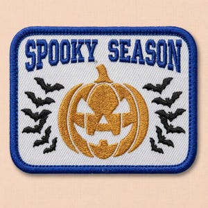 Pode incluir: Um emblema retangular bordado com fundo branco e borda azul. O texto SPOOKY SEASON está bordado em azul no topo. Uma abóbora dourada fica no centro, ladeada por pequenos morcegos pretos bordados em ambos os lados da peça.