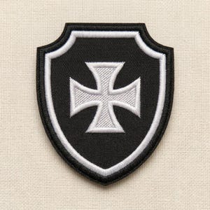 Puede incluir: Un parche bordado en forma de escudo que presenta un fondo negro con un borde blanco y un emblema de cruz blanca centrado. El diseño está cosido con una textura de hilo densa, creando una apariencia en relieve sobre la superficie de la tela.