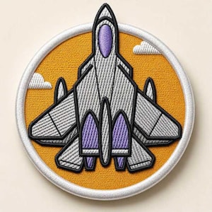 May include: A circular embroidered patch featuring a gray fighter jet with purple accents on a mustard yellow background. The design includes two small white clouds and is framed by a thick white border, showcasing detailed stitching throughout.