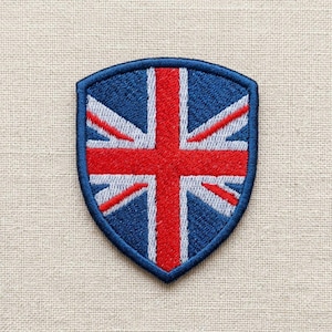 Puede incluir: Un parche bordado en forma de escudo con el diseño de la bandera Union Jack. El parche tiene un borde azul oscuro y muestra la cruz y las franjas diagonales rojas, blancas y azules de la bandera británica con un acabado de hilo texturizado.