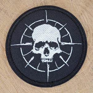 May include: A circular embroidered patch featuring a white skull centered within a white crosshair target design on a black background. The patch has a thick black stitched border and a textured fabric surface with detailed white thread embroidery.