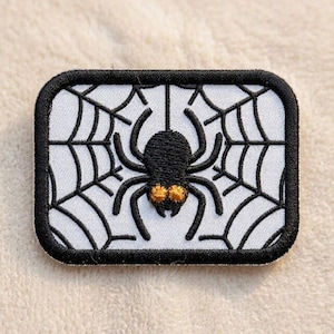 May include: A rectangular embroidered patch featuring a black spider centered on a white spiderweb background. The spider has eight legs and two prominent golden-yellow eyes. The entire patch is framed by a thick, black stitched border.