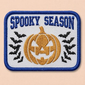 Puede incluir: Un parche rectangular bordado con fondo blanco y borde azul. El texto SPOOKY SEASON está bordado en azul en la parte superior. Una calabaza dorada se sitúa en el centro, flanqueada por pequeños murciélagos negros bordados a ambos lados.