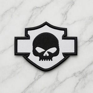 Könnte beinhalten: Ein schildförmiger Stickaufnäher mit einer schwarzen Totenkopfgrafik auf weißem Hintergrund. Der Aufnäher hat einen dicken schwarzen Rand und ein markantes horizontales Balkendesign, das sich zu den Seiten erstreckt und eine symmetrische Form bildet.