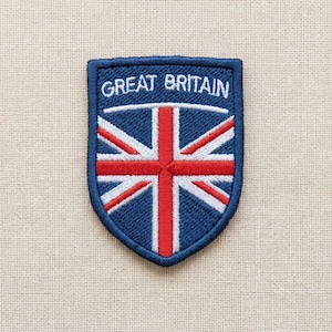 Op de afbeelding: Een schildvormige geborduurde patch met de Union Jack-vlag in rood, wit en blauw. De tekst GREAT BRITAIN is in witte hoofdletters geborduurd op het bovenste gedeelte van de blauwe achtergrond, omrand door een donkerblauwe rand.