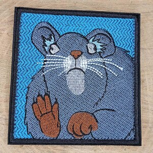 May include: A square embroidered patch featuring a dark gray rodent with a light gray belly, brown paws, and white whiskers. The rodent is set against a bright blue background with a chevron pattern, all enclosed by a thick black border.