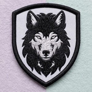 Puede incluir: Un parche bordado en forma de escudo que presenta una cabeza de lobo detallada en blanco y negro. El lobo tiene orejas puntiagudas, ojos enfocados y un patrón de pelaje texturizado. El diseño está rodeado por un borde negro grueso sobre fondo blanco.