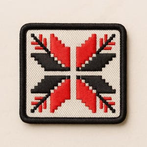 Könnte beinhalten: Ein quadratischer Stickaufnäher mit einem geometrischen, pixelartigen, blumenähnlichen Muster aus rotem und schwarzem Garn auf cremefarbenem Stoffgrund. Das Design ist symmetrisch und von einem dicken, schwarzen Rand umgeben.