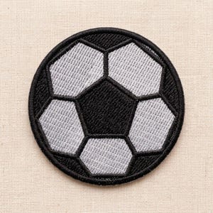 Puede incluir: Un parche bordado circular con un diseño clásico de balón de fútbol. El patrón consiste en formas pentagonales y hexagonales alternas en negro y gris claro, todas delineadas con costuras de hilo negro grueso en una forma redonda definida.