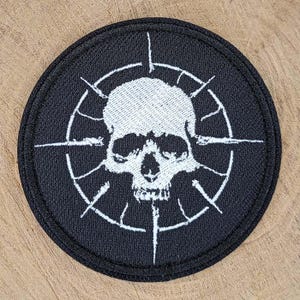 May include: A circular embroidered patch featuring a white skull centred within a white circular frame with radiating lines. The background of the patch is black, and the entire design is stitched with a thick black border around the outer edge.