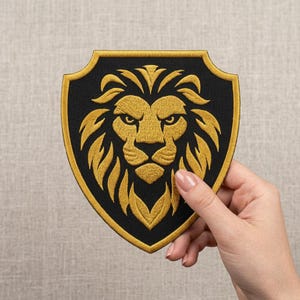 May include: A shield-shaped embroidered patch featuring a detailed golden lion head centered on a solid black background. The patch is outlined with a thick golden border, creating a sharp contrast against the black fabric and the intricate lion mane.