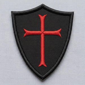 Puede incluir: Un parche bordado en forma de escudo con fondo negro y un borde negro grueso. Centrada en el parche hay una cruz roja con extremos ensanchados, cosida con una textura de hilo visible que crea una apariencia detallada y en relieve.