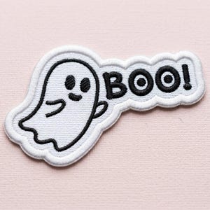 Puede incluir: Un parche bordado blanco que muestra un fantasma de dibujos animados con contornos y rasgos faciales negros. A la derecha del fantasma, la palabra BOO! está bordada en letras negras negritas. El diseño tiene un borde blanco y acabado texturizado.