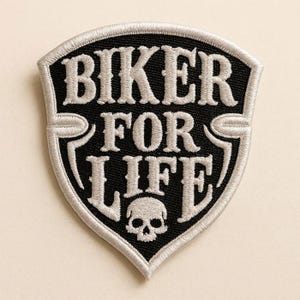 May include: A shield-shaped embroidered patch with a black background and silver-colored border. The text BIKER FOR LIFE is stitched in silver thread in a bold font. A small silver skull icon is positioned at the bottom center of the patch.