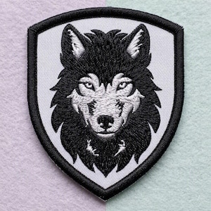 Puede incluir: Un parche bordado en forma de escudo que presenta una cabeza de lobo detallada en blanco y negro. El lobo tiene orejas puntiagudas, ojos enfocados y un patrón de pelaje texturizado. El diseño está rodeado por un borde negro grueso sobre fondo blanco.